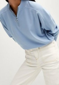 Sudadera azul claro con cuello de cremallera parcial y tirador circular, combinada con vaqueros blancos de cintura alta con bolsillos traseros y detalles cosidos.