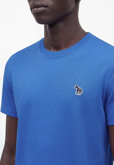 Άνδρας που φοράει έναν μονόχρωμο μπλε T-shirt με ένα μικρό πολύχρωμο έμβλημα ζέβρας στο αριστερό στήθος.