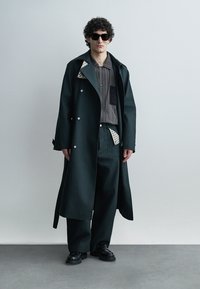 Homme portant des lunettes de soleil noires, un long manteau sombre sur une chemise rayée avec un patch, un pantalon large noir et des chaussures noires, debout devant un fond uni.