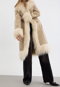 Cappotto in pelliccia ecologica beige con maniche lunghe, polsini texturizzati e colletto. Presenta una vita con cintura e dettagli di cucitura a contrasto. Indossato su pantaloni neri.