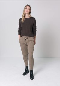 Vrouw staand in een casual outfit met een donker longsleeve trui, beige broek met trekkoord, zwarte enkellaarsjes, tegen een effen lichte achtergrond.