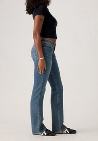 Femme debout de profil portant un jean évasé bleu, un t-shirt court noir, des baskets noires et blanches, et un bracelet argenté au poignet gauche.