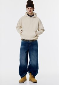 Beige oversized mikina s predným vreckom, modré široké nohavice a béžové boots. Hnedá pletená čiapka dopĺňa uvoľnený vzhľad.