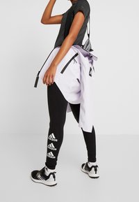 Persona vistiendo leggings y zapatillas negras de Adidas, sosteniendo una chaqueta morada clara sobre su hombro contra un fondo liso.