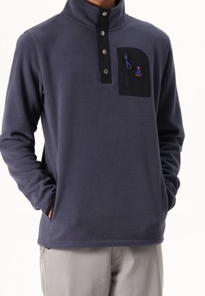 Person iført en mørkeblå fleece-pullover med sort trykknap-åbning og en lynlåslomme på brystet, hænder i sidelommer.