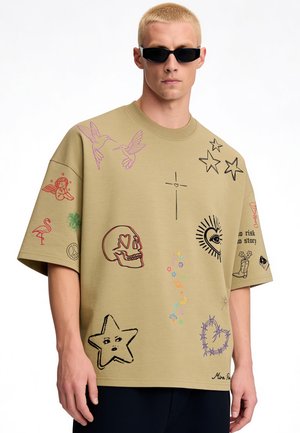 Homme portant un t-shirt oversize beige avec des motifs colorés de style tatouage comprenant un crâne, une étoile, des colibris, un cœur et du texte sur les manches courtes et à l'avant.