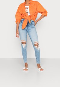 Camisa naranja de botones atada a la cintura, blusa blanca con estampado debajo, jeans ajustados rotos de color azul claro y sandalias blancas.