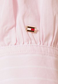 Roze stof met een geplooide textuur, voorzien van een rechthoekige gouden hardware-accent met rode, witte en zwarte details.