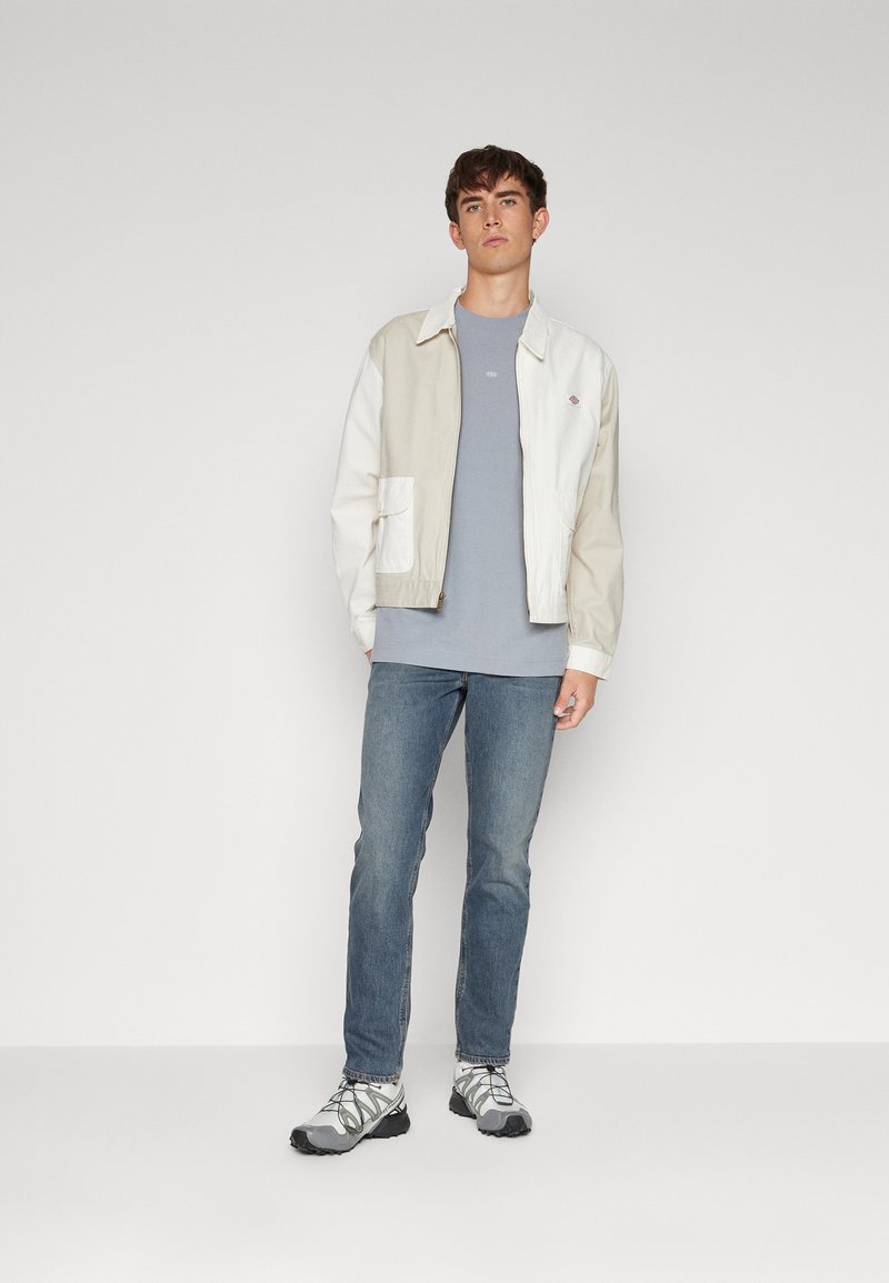 Lichtgewicht, twee-kleurige jack in crème en beige, gecombineerd met een blauwe T-shirt, blauwe jeans en grijze sportschoenen. Beschikt over een voorzak.