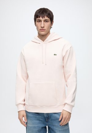 Giovane uomo che indossa una felpa con cappuccio rosa chiaro con tasca a marsupio e piccolo logo verde sul petto, abbinata a jeans blu, in piedi davanti a sfondo bianco.