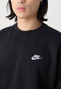 Personne portant un sweat-shirt Nike noir avec logo blanc, cheveux courts noirs et un anneau d'oreille argenté sur un fond uni.