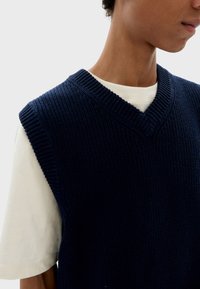 Gilet tricoté bleu marine avec un col en V, présentant une texture côtelée et un design sans manches, superposé sur un t-shirt blanc à col rond.