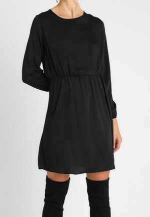 Schwarzes langärmeliges Kleid mit Rundhalsausschnitt und elastischer Taille, gefertigt aus leichtem Stoff, knielang, kombiniert mit hohen schwarzen Stiefeln.