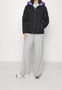 Zwarte gewatteerde jas met een paarse capuchon, rits aan de voorkant en zijzakken, gecombineerd met lichtgrijze wijde broek en grijze sneakers.