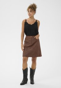 Camisole noire avec des détails en dentelle, associée à une jupe en cuir synthétique marron avec une poche avant et une fermeture à bouton. Portée avec des bottes de cowboy noires.