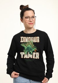 Felpa nera con una stampa grafica di un dinosauro e la scritta "DOMATORE DI DINOSAURI" in grassetto, colori arancione e verde. Materiale in cotone morbido.