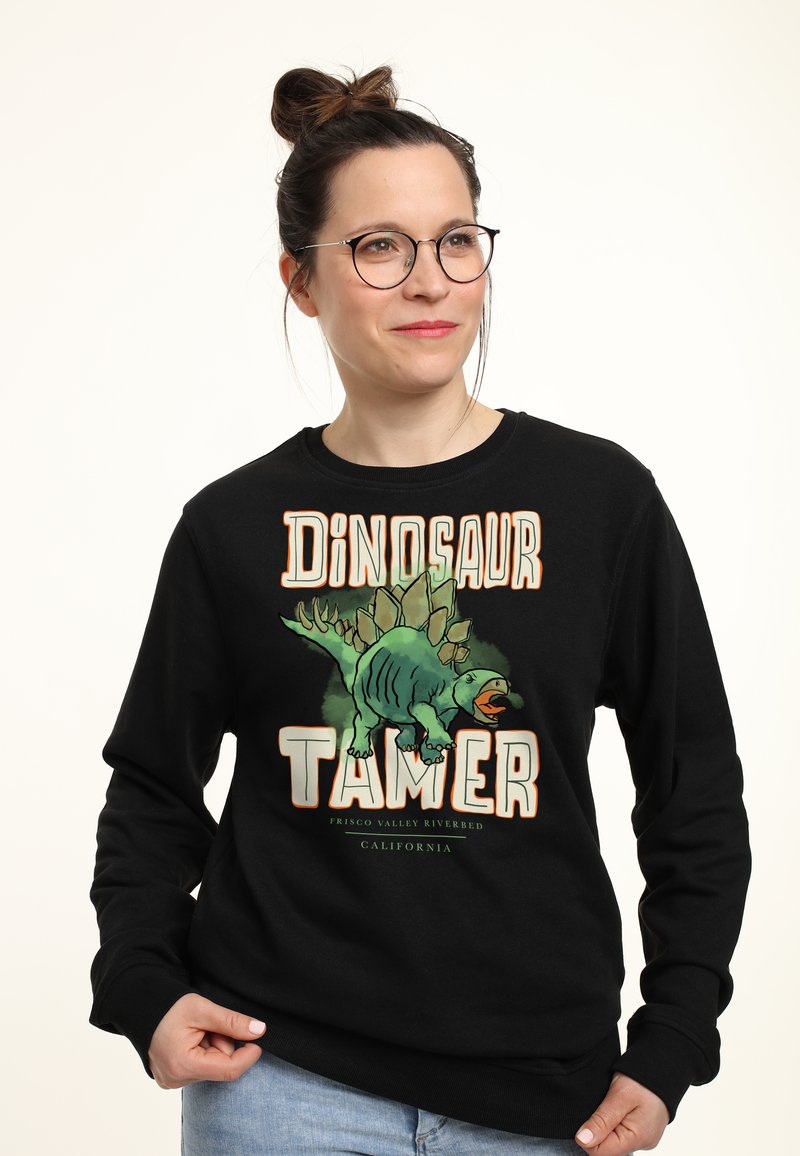 Felpa nera con una stampa grafica di un dinosauro e la scritta "DOMATORE DI DINOSAURI" in grassetto, colori arancione e verde. Materiale in cotone morbido.