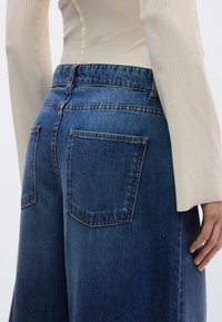 Τζιν παντελόνια με φαρδιά πόδια σε σκούρο μπλε χρώμα, με δύο πίσω τσέπες και ορατές ραφές. Ζώνη υψηλής μέσης με ελαστική λεπτομέρεια. Απαλή υφή.