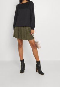 Haut noir à manches longues, jupe en suède vert olive avec poches, bottines à talons noires et sac à main beige clair. Texture lisse et style superposé.