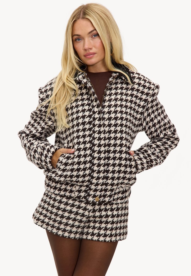 Femme blonde portant une veste et un short assortis en pied-de-poule noir et blanc, les mains dans les poches de la veste, regardant l'appareil photo.