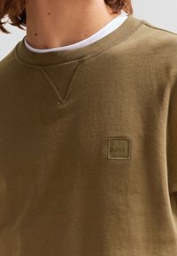 Felpa verde oliva con scollo a girocollo, caratterizzata da una discreta toppa con il logo BOSS. Tessuto morbido e vestibilità leggermente comoda con dettagli delle cuciture.