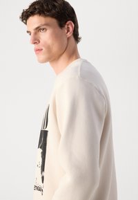 Young man with short curly hair wearing an off-white sweatshirt with a black graphic, looking to the left against a plain light background.