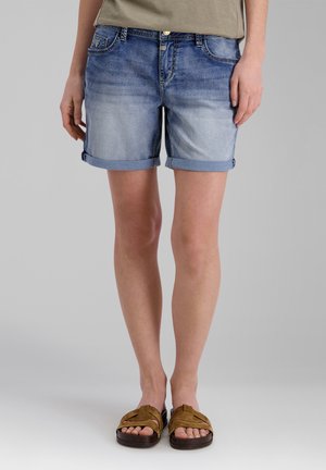 Erwachsener trägt hellblaue hochgekrempelte Jeans-Shorts, olivgrünes Oberteil und braune Sandalen auf schlichtem grauem Hintergrund.
