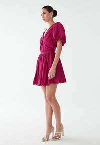 Fuchsias Kleid mit Puffärmeln, einem tiefen V-Ausschnitt und einem schwingenden Rock. Verfügt über eine geraffte Taille und einen gewellten Saum. Trägt weiße Absatzsandalen.