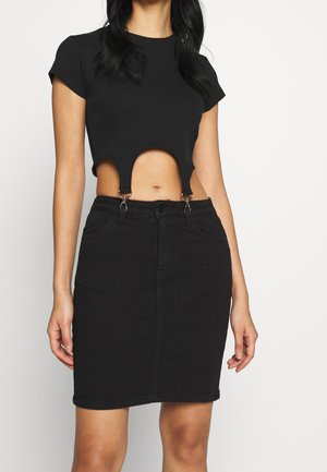 Woman wearing a black cropped top with cutouts and metal clasps paired with a high-waisted black denim skirt against a white background.