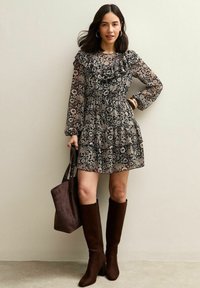 Zwarte bloemenjurk met lange doorschijnende mouwen, gerimpelde halslijn en een rokje met meerdere lagen. Gecombineerd met bruine kniehoge laarzen en een bruine shopper.