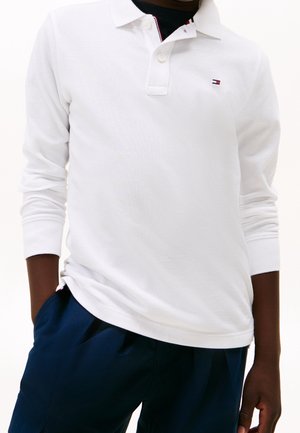 Homme portant un polo blanc à manches longues avec un petit logo et un pantalon bleu marine, les mains partiellement dans les poches.