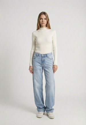 Femme debout, face à l'avant, portant un haut blanc côtelé à manches longues, un jean ample bleu clair et des baskets blanches sur un fond uni.