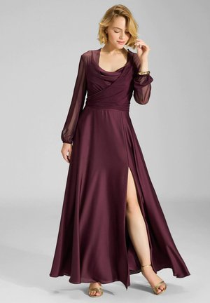 Donna che indossa un lungo vestito bordeaux con maniche lunghe trasparenti, spacco laterale e sandali dorati, posando con una mano vicino al viso.