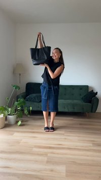 Black leather tote bag with two long handles, smooth texture, and a rounded shape, held by a person in a casual outfit against a neutral backdrop.