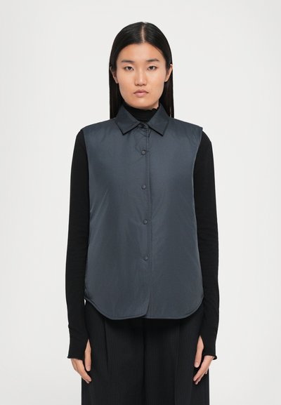 Donkergrijs mouwloos vest met een knoopsluiting aan de voorkant, kraag en gebogen zoom. Gemaakt om te dragen over een zwart longsleeve top. Gladde stoftextuur.