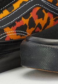 Gros plan sur deux baskets montrant des semelles en caoutchouc noires, un bout en daim noir, et une tige en tissu avec un imprimé léopard orange, rouge et noir ainsi que des coutures blanches.