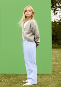 Grå stickad cardigan över en vit skjorta, kombinerad med ljusblå randiga byxor. Outfitten kompletteras av vita skor. Grön bakgrund.