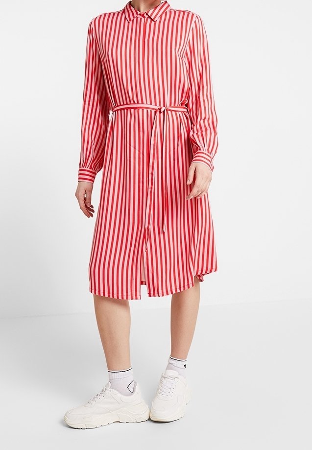 Femme portant une robe à manches longues rayée verticalement rouge et blanche avec une ceinture à la taille, des chaussettes blanches et des baskets blanches épaisses, debout sur un fond uni.