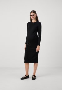 Vestido de maternidade preto de manga longa, corpete justo, até ao joelho, tecido liso, usado com sapatos pretos planos, modelo de pé contra um fundo neutro.