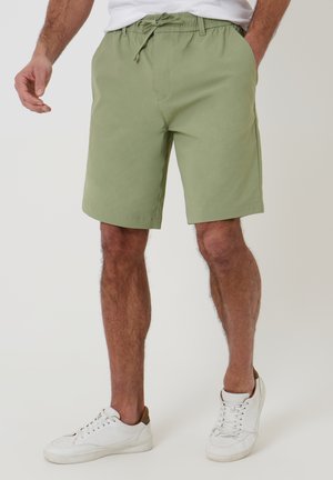 Mand iført lysegrønne knælange shorts med elastisk talje og hvide sneakers, stående mod en ensfarvet baggrund.