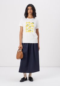 Weißes Baumwoll-T-Shirt mit gelbem Zitronenprint, kombiniert mit einem langen marineblauen Rock. Eine kleine gewebte braune Handtasche rundet das Outfit ab. Schwarze Schuhe und beige Socken.