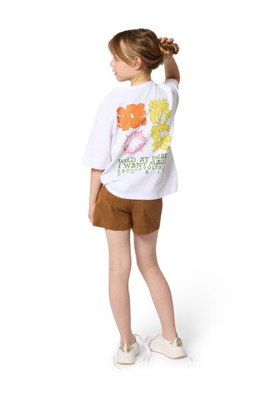 Ragazza giovane che sistema lo chignon, indossa una maglietta bianca con stampe floreali colorate e testi, pantaloncini marroni e sneakers bianche con dettagli dorati.