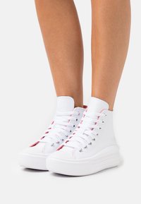 Nogi w białych wysokich trampkach z grubą podeszwą i białymi sznurowadłami na jednolitym jasnym tle.