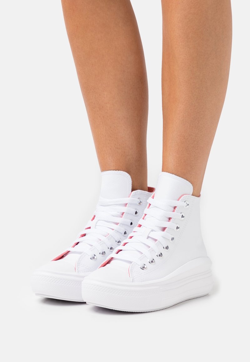 Nogi w białych wysokich trampkach z grubą podeszwą i białymi sznurowadłami na jednolitym jasnym tle.