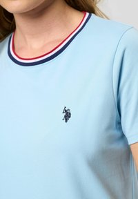 Svijetloplava pamučna majica s okruglim izrezom, s crvenim i tamnoplavim prugama te malim tamnim izvezenim logom na prsima.