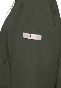 Ciemnozielona kurtka z białą materiałową etykietą, na której znajduje się czarne logo psa oraz różowy napis „FREDDY FREDDIES NEW YORK”.