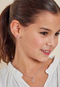 Silver cross-halsband med rosa detaljer, bärs över en vit texturerad blus. Små örhängen med rosa och klara stenar.