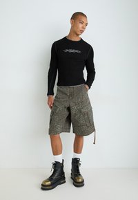 Schwarzes Langarmshirt mit silberner Grafik, kombiniert mit grauen Cargo-Shorts im Leopardenmuster und schwarzen Stiefeln mit goldenen Akzenten.