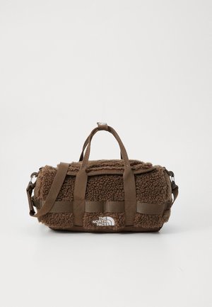 Sac de sport marron en tissu texturé et moelleux, avec des sangles tissées, un patch logo et une fermeture éclair. Il dispose d'une bandoulière amovible.