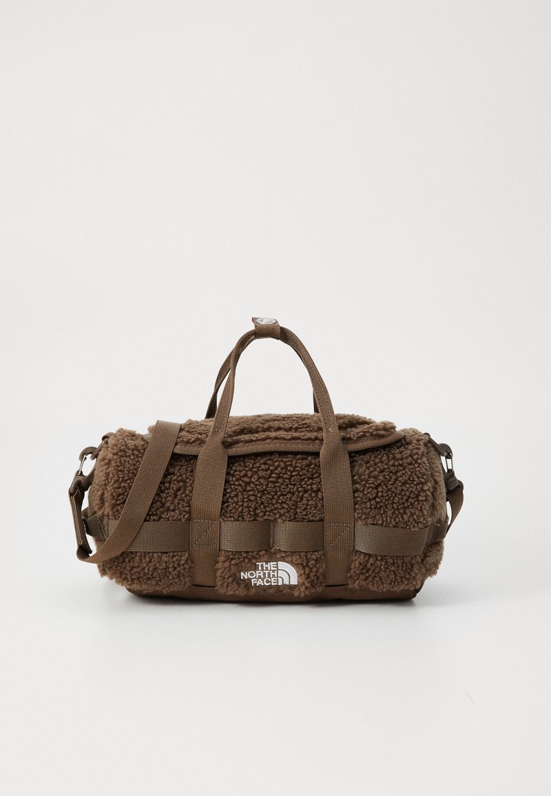 Brown duffel bag made of textured, fluffy fabric with webbed straps, logo patch, and a zip closure. Features a removable shoulder strap.